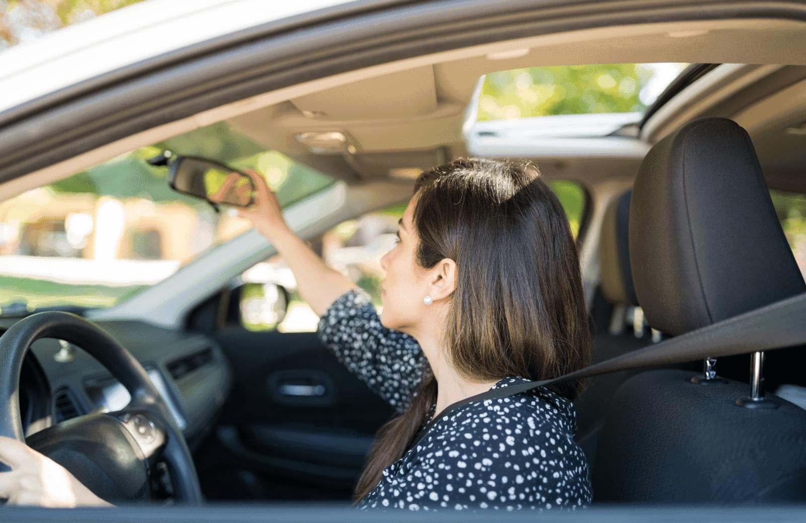 A woman sits in the driver seat of a car and adjusts the review mirror