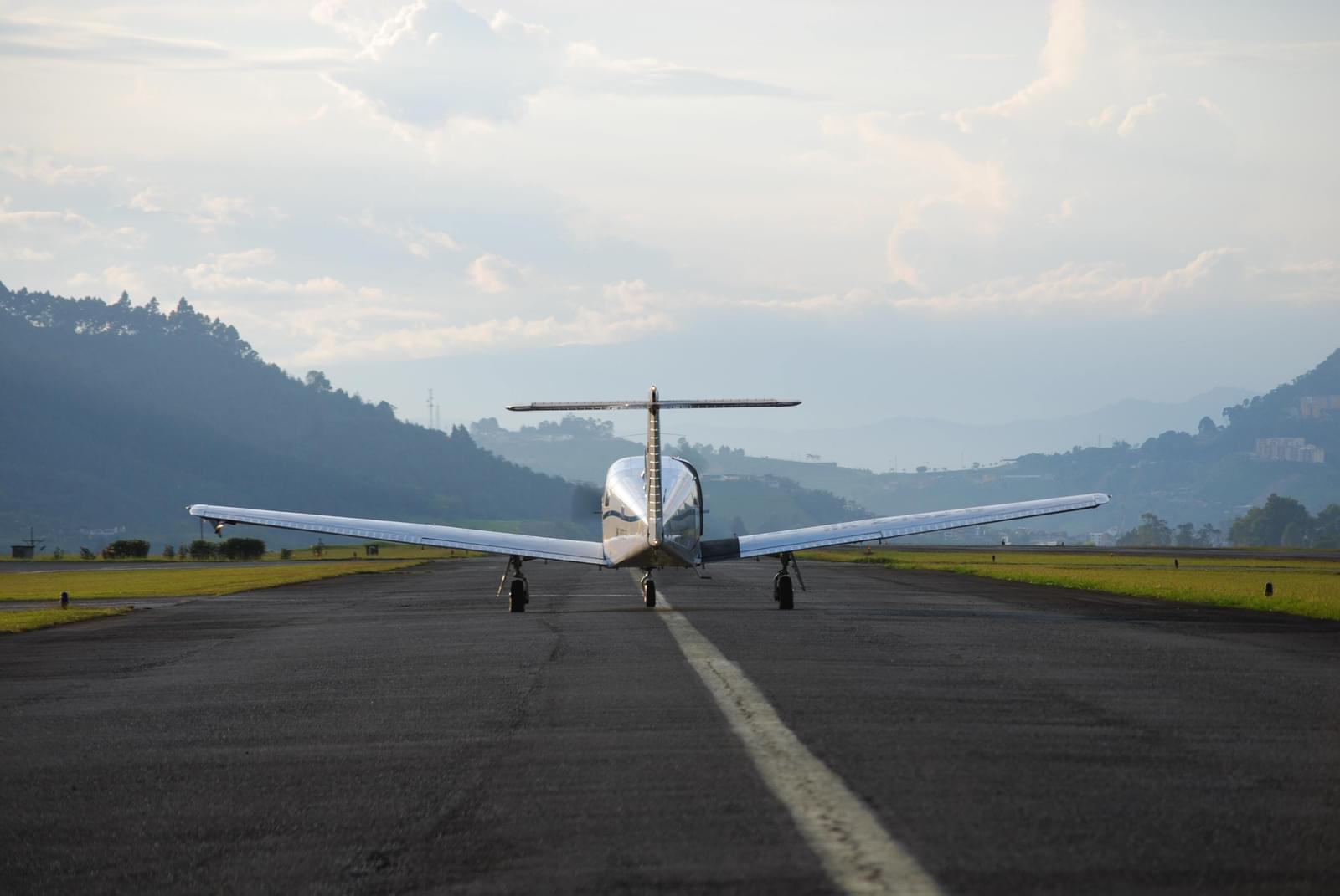 A shiny chrome airplane gets ready to launch into flight on a runway. The runway is surrounded by bright green grass and a beautiful mountainscape is in the background, in the direction the plane is taking off into.