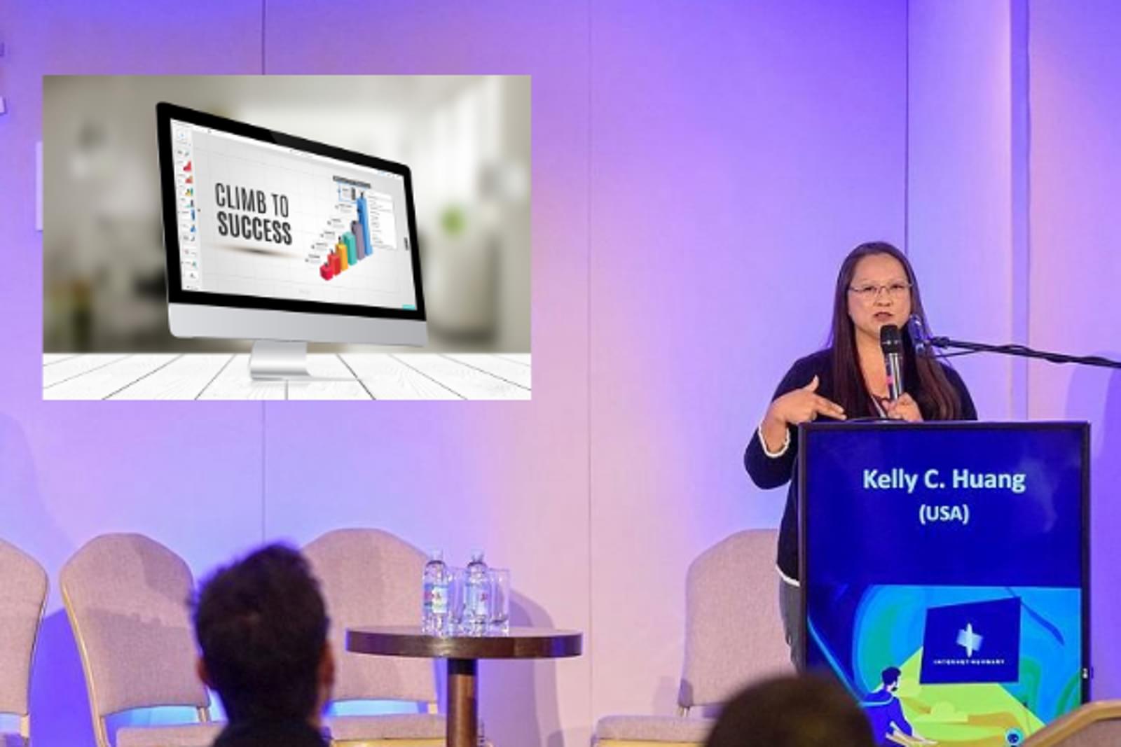 Kelly Huang stands behind a podium in a Conference setting. She is speaking into a microphone and gesturing as she presents to the Conference audience. A screen projected on the Conference wall behind her shows an image and the text "Climb to Success."