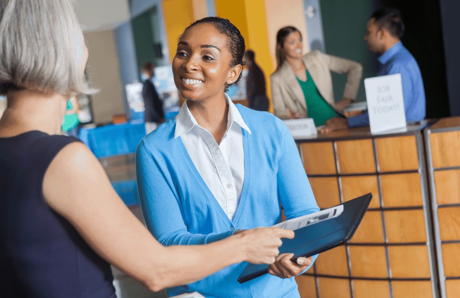 A woman wearing a white dress shirt with a blue sweater shirt smiles as at another woman, dressed in a dark blue shirt. The woman in a dark blue shirt is handing her resume to the woman in the white shirt at a job fair.