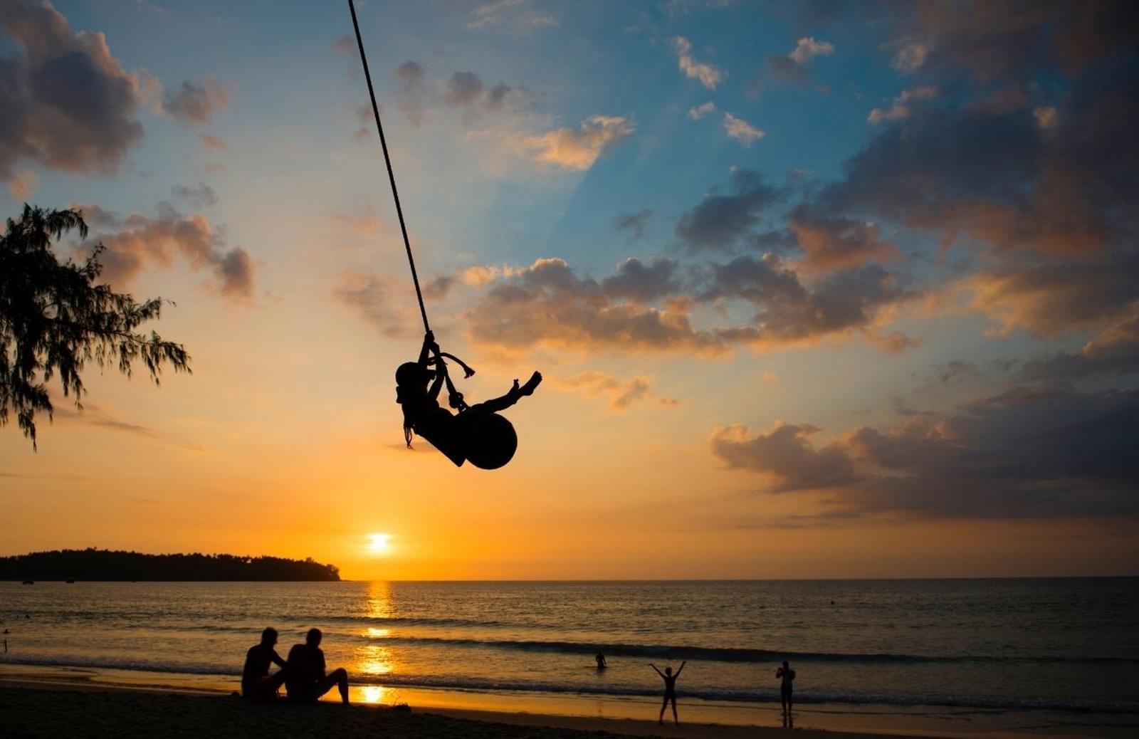 A summer sun sets over a body of water. Silhouettes of a family, lounging, playing and swinging on a rope are on the shoreline.