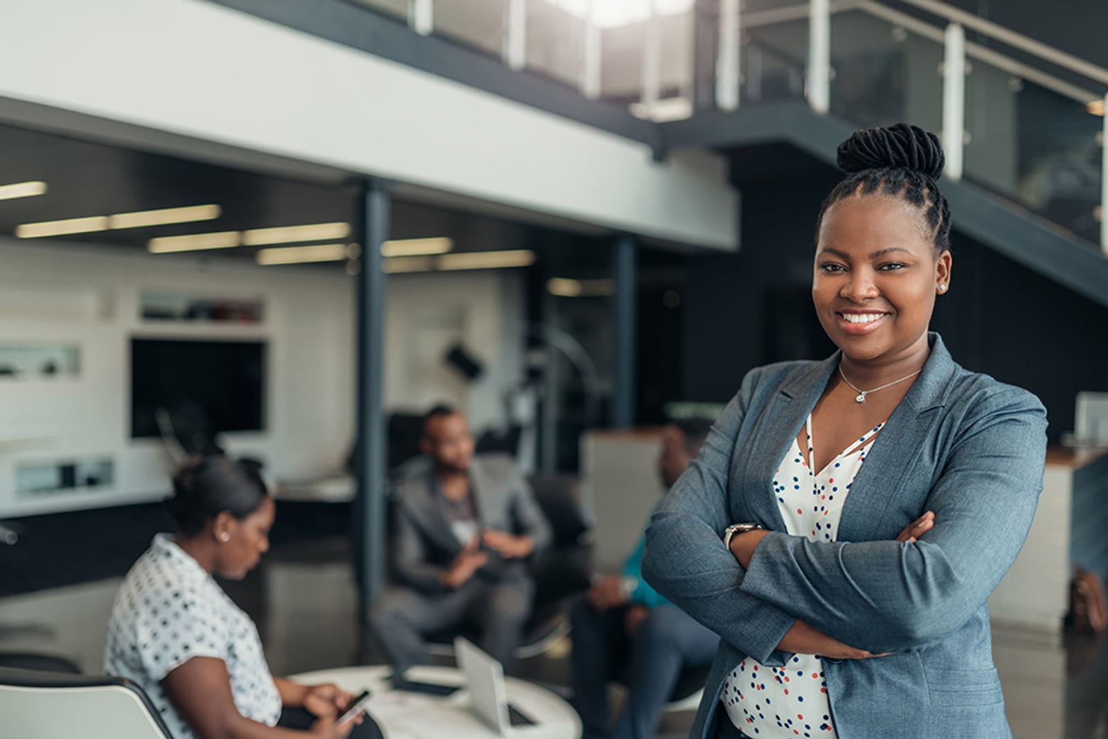 Confident business woman crosses arms smiling at camera in office with coworkers behind her