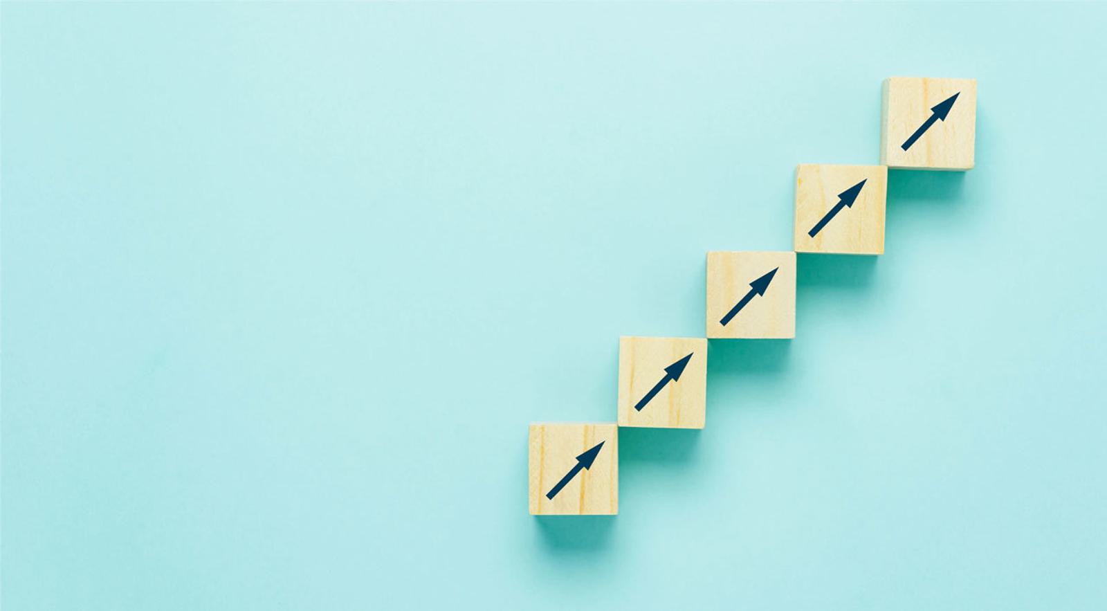 Five wooden blocks with upward facing arrows on incline with light blue background