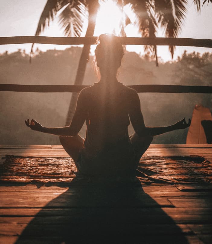 The sun sets behind a palm tree showing a silhouette of a woman in a meditative pose with her arms out to the side