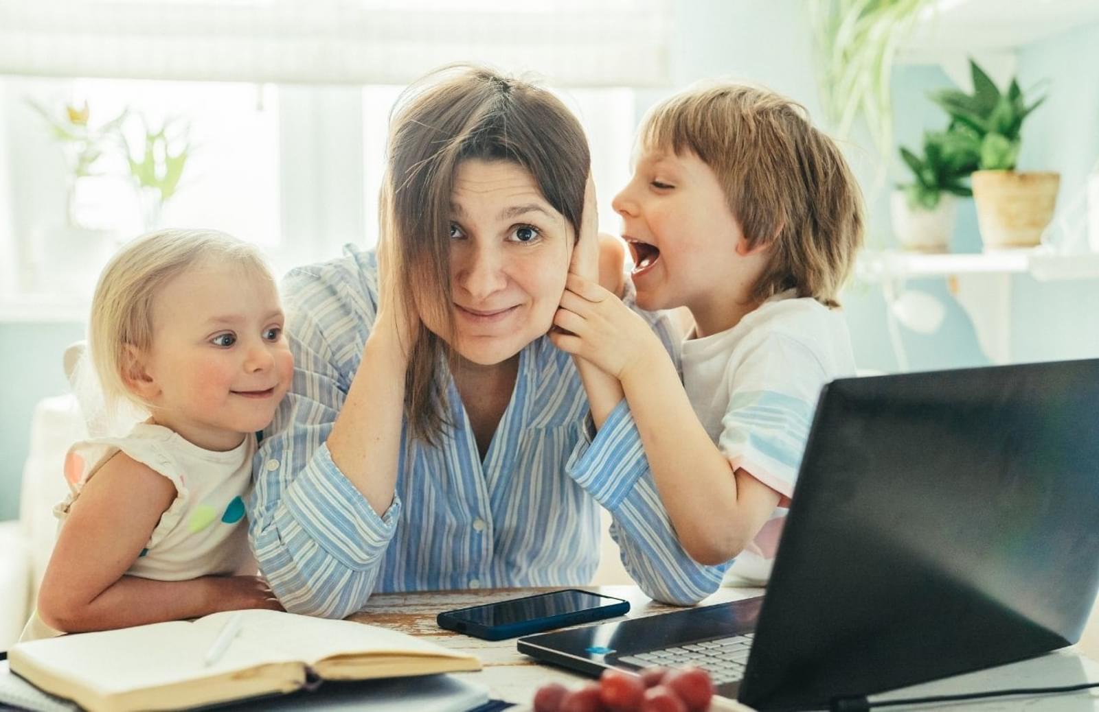 Woman at desk with hands over her ears while young children talk to her