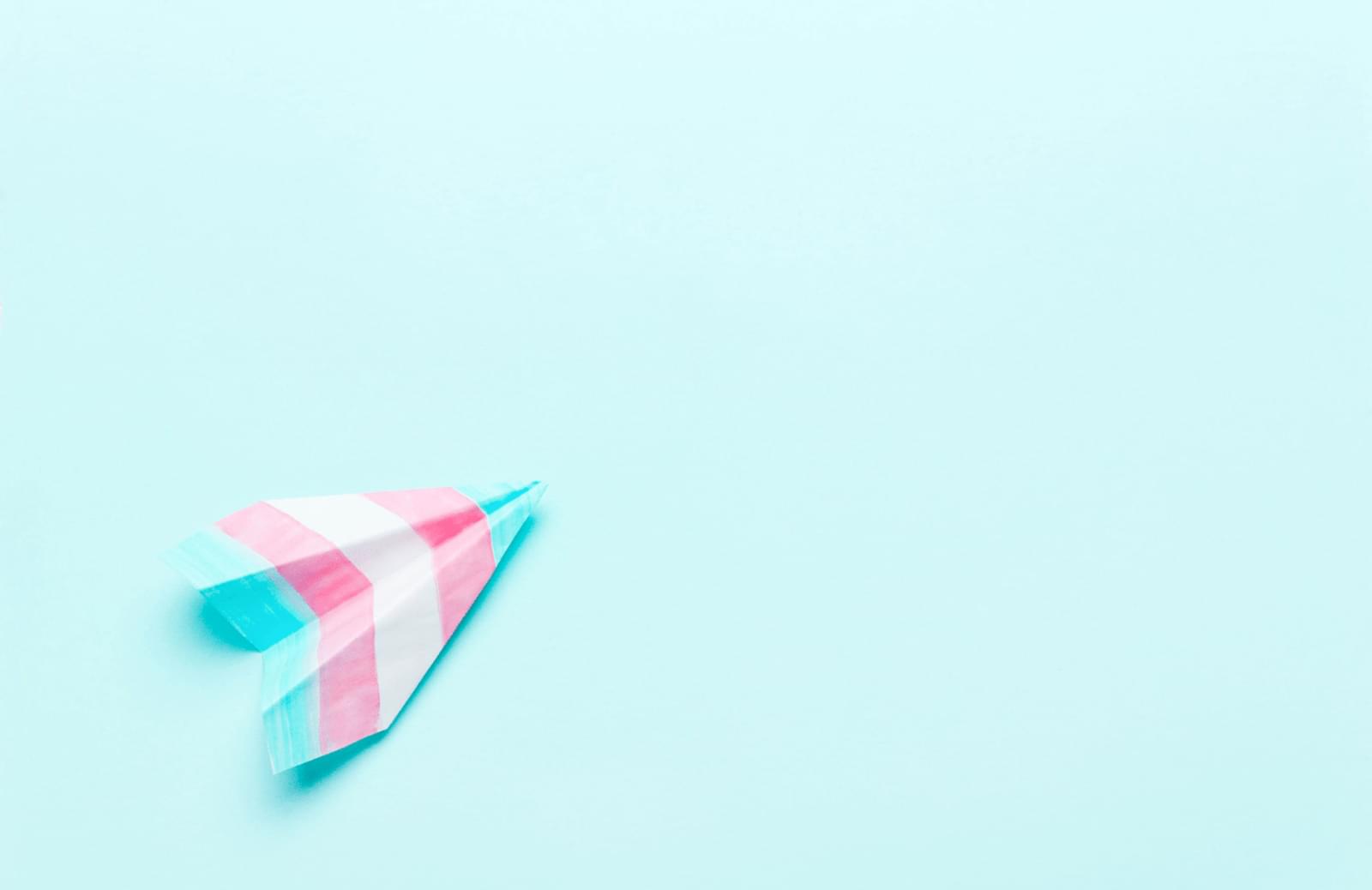 A paper airplane with the colors of the transgender flag on it is folded and pointed upright on a plain teal background