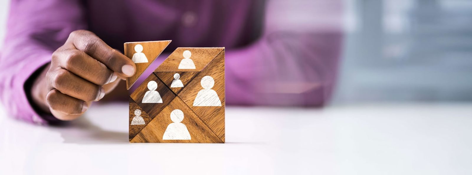 Someone wearing a purple dress shirt builds a wooden tanagram square block with icons of people painted on each puzzle piece