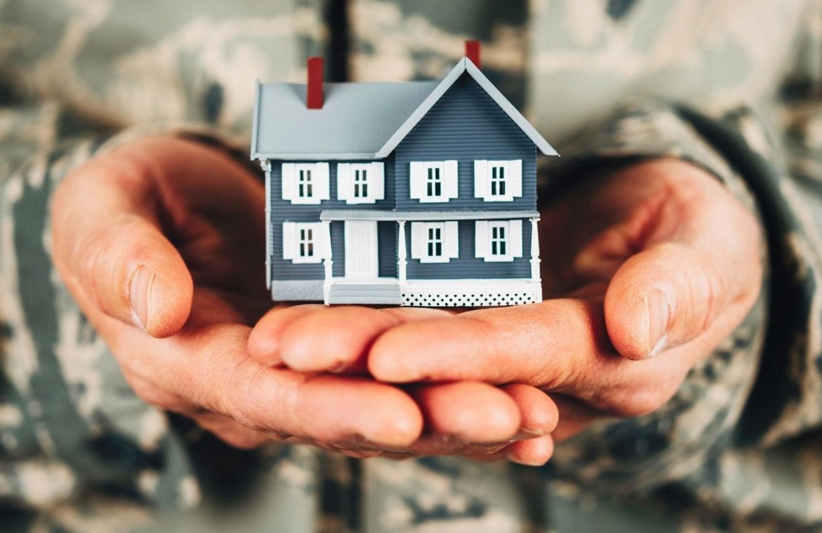 Person dressed in  military fatigues holding tiny house replica in palms
