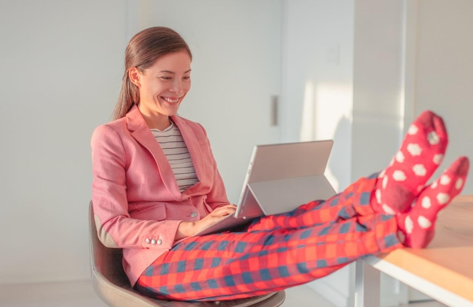 Woman with feet on desk, wearing pajamas, smiling while working on laptop computer