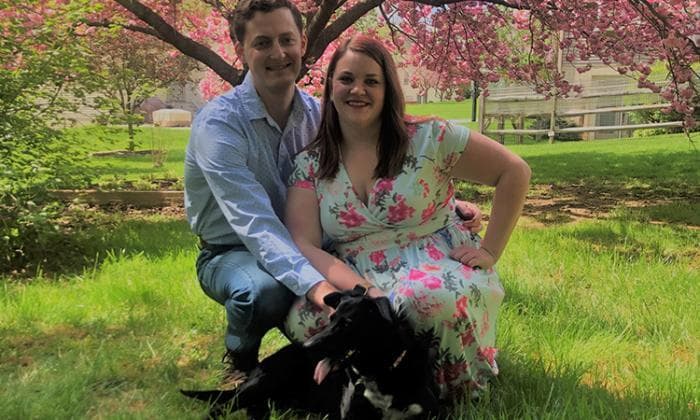 Shannon and her husband pose with their black dog, Bailey. A blossoming cherry tree is behind them and lots of bright green grass.