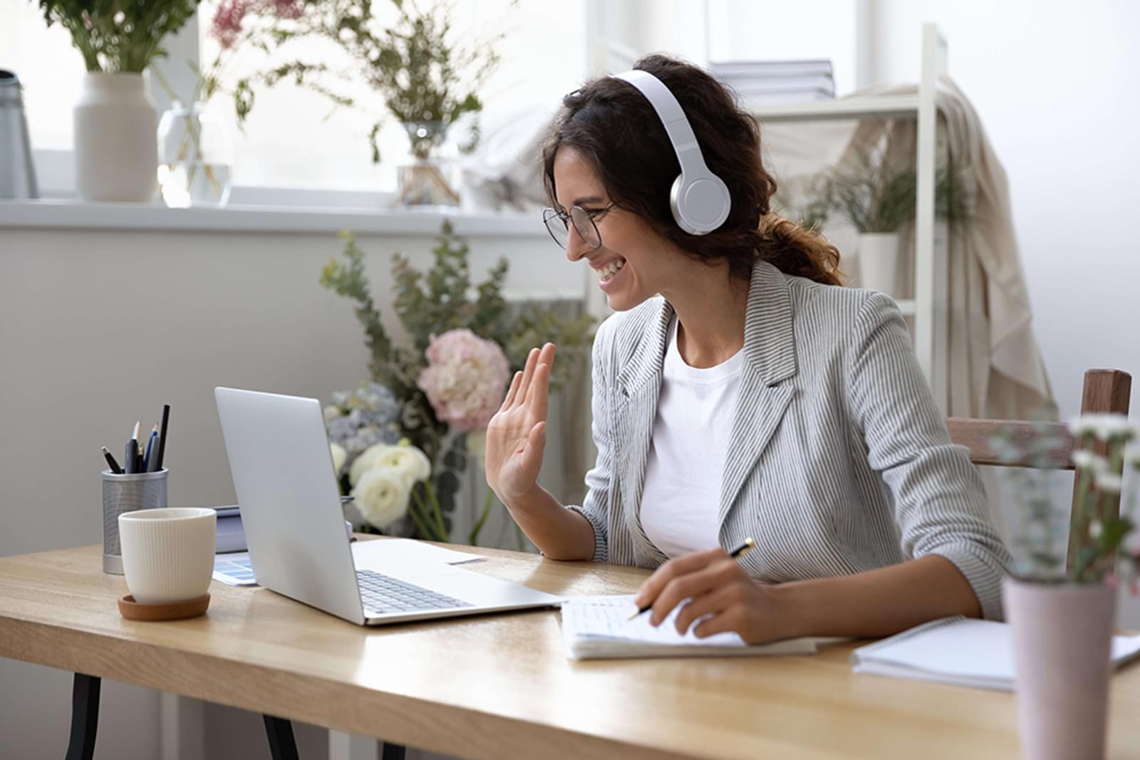 Smiling woman attending webinar wears headset waves at computer screen
