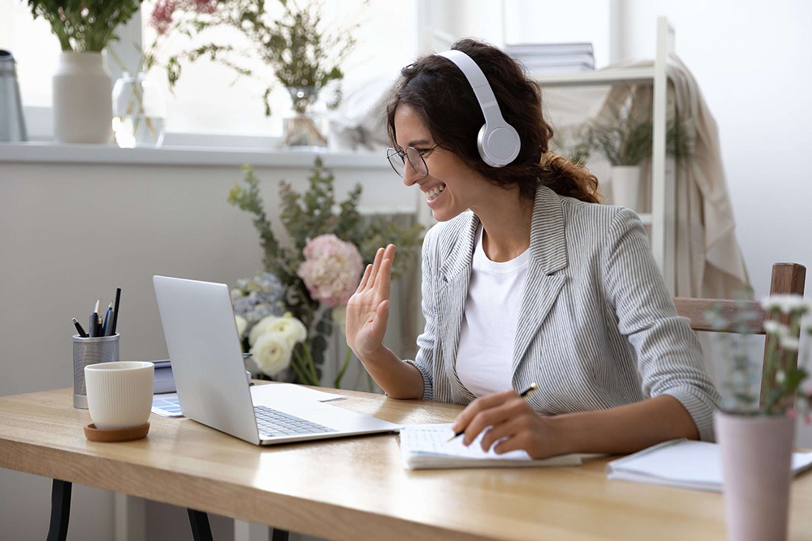 Smiling woman attending webinar wears headset waves at computer screen