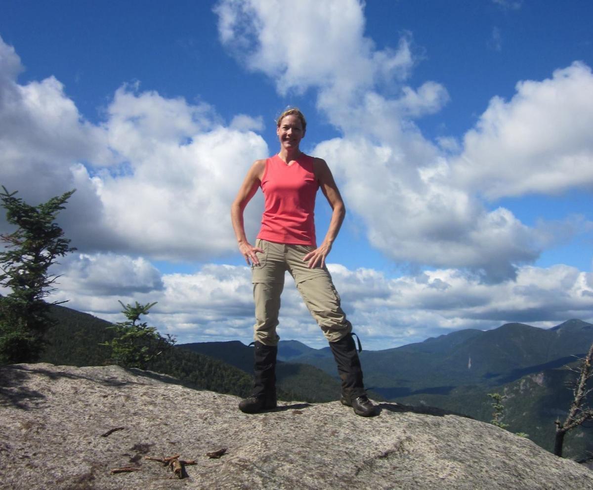 Jennifer Howland with her hands on her hips with a blue sky and white clouds behind her as she poses on a mountain during hiking with a mountain range in the backdrop