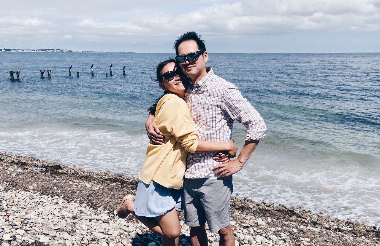 Kellie and her husband on a rocky beach with the ocean and clear blue skies behind them