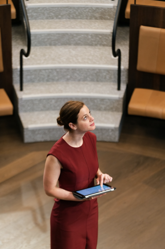 A woman standing in a lecture hall looks upward off camera as she adjusts the settings with her finger on a tablet. She is wearing a red dress and has her hair pulled back in a bun.